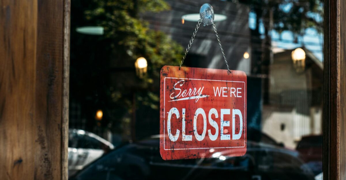 A red closed sign hangs on a glass door reflecting cars and trees outside