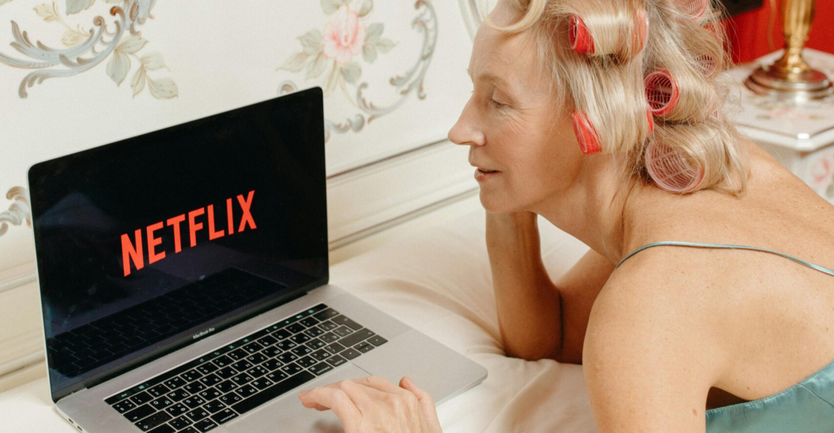Senior woman with curlers enjoying Netflix on a laptop while lying in a stylish bedroom.
