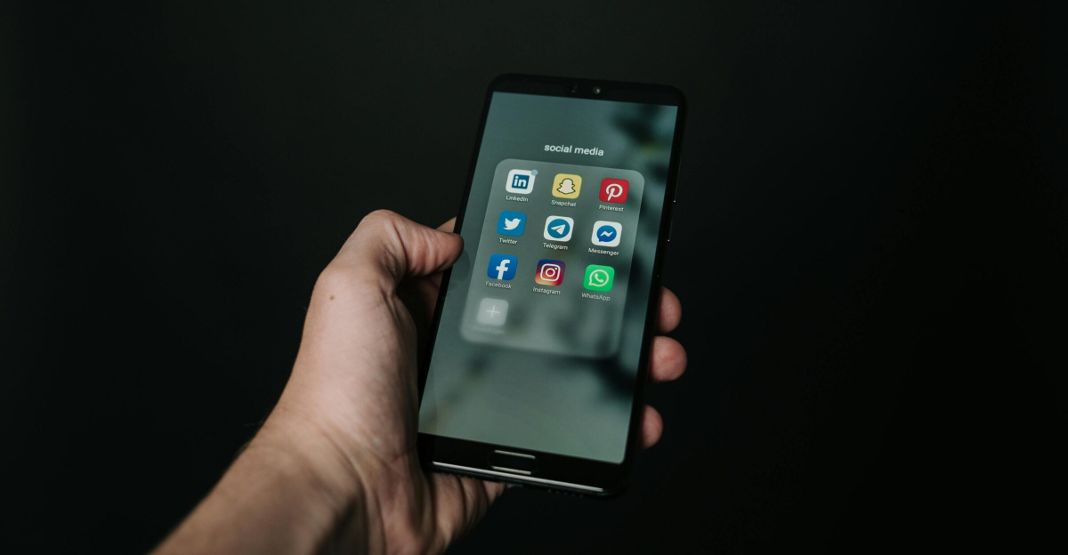 Close-up of a hand holding a smartphone displaying various social media app icons on a dark background.