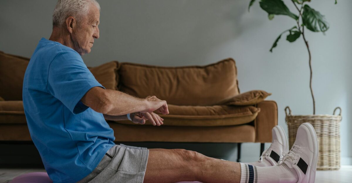 Elderly man in blue shirt exercising on a yoga mat indoors with dumbbells in view