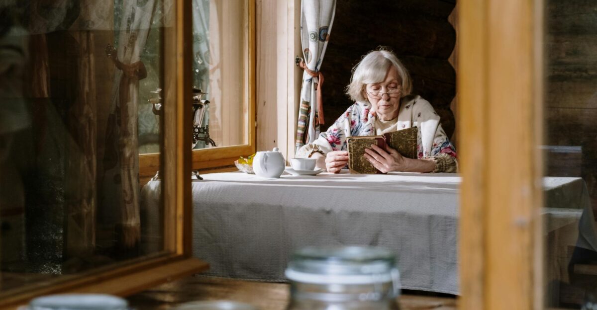 Senior woman reads by the window in a cozy cabin enjoying a cup of tea with a teapot on the table